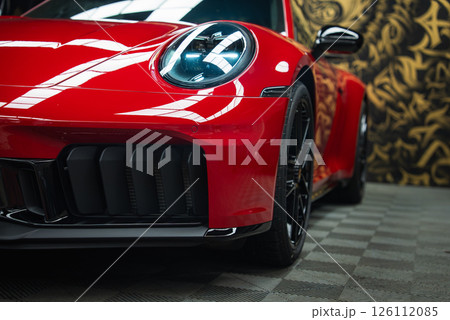 Front end of a red 2025 Porsche 911 GT3 922 with LED headlights, black alloy wheels, and a glossy finish. Ornate gold and black wall in the background. 126112085