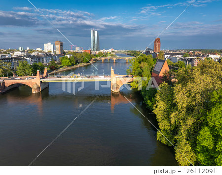 Aerial view of Frankfurt, Germany, featuring the Main River, Alte Brucke, European Central Bank tower, green trees, and a red roofed building under a partly cloudy sky. 126112091