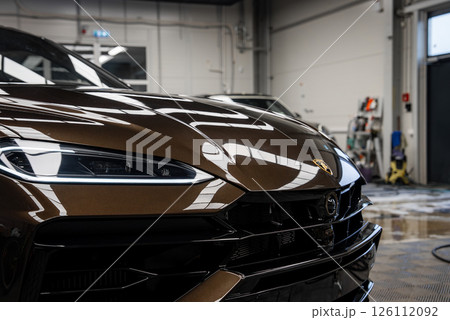 Front end of a metallic brown Lamborghini Urus with sharp LED headlight and emblem, set in a garage with tools, hose, and cleaning supplies visible. Front end of a metallic brown Lamborghini Urus with sharp LED headlight and emblem, set in a garage with tools, hose, and cleaning supplies visible. 126112092