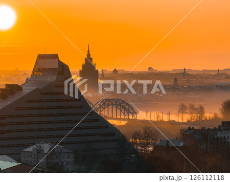 A sunrise view of Riga, Latvia, showcasing the National Library, Riga TV Tower, Daugava River, and arched railway bridge under a misty orange sky. 126112118
