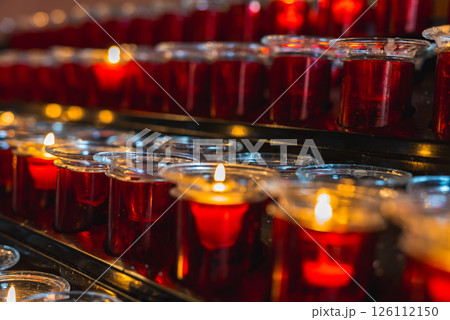 Red votive candles in glass holders, some lit and others unlit, arranged on a black metal stand in a serene religious or spiritual setting. 126112150