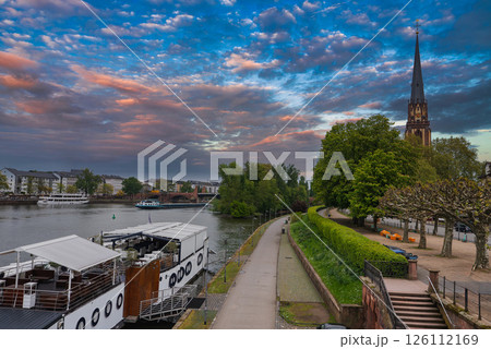 View of Frankfurt, Germany, featuring the Main River, a docked boat, Dreikonigskirche spire, a tree lined pathway, and a mix of urban architecture. View of Frankfurt, Germany, featuring the Main River, a docked boat, Dreikonigskirche spire, a tree lined pathway, and a mix of urban architecture. 126112169