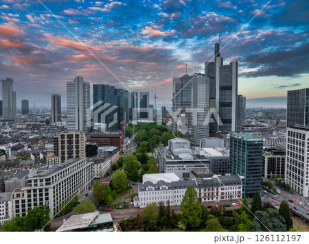 Aerial view of Frankfurt, Germany, featuring modern skyscrapers like Commerzbank Tower, historic buildings, green spaces, and a vibrant sunset sky. 126112197