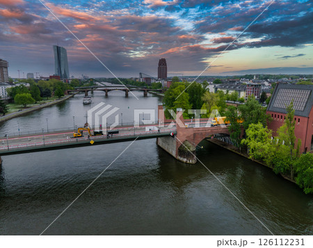 Aerial image of Frankfurt, Germany, showing the Main River, a bridge with construction vehicles, the ECB building, and a red brick tower at sunset. Aerial image of Frankfurt, Germany, showing the Main River, a bridge with construction vehicles, the ECB building, and a red brick tower at sunset. 126112231