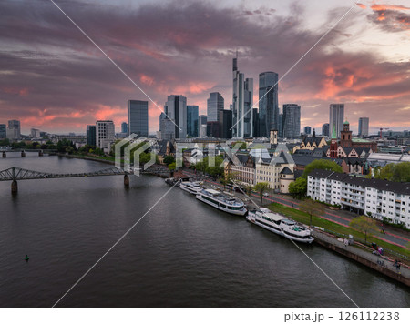 Aerial view of Frankfurt, Germany, featuring the Commerzbank Tower, modern skyscrapers, the Main River, a pedestrian bridge, and a vibrant sunset. 126112238