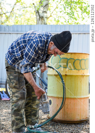 Elderly Russian man standing by a barrel with a dry pump in his hands, checking the hose before work in a quiet rural yard 126112302