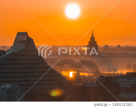 The image shows a sunrise over Riga, Latvia, featuring the National Library, the Latvian Academy of Sciences, the Daugava River, and a railway bridge. 126112362