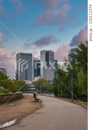 A park pathway with greenery and a bench leads to modern skyscrapers in Frankfurt, Germany. The overcast sky highlights the urban skyline. 126112374