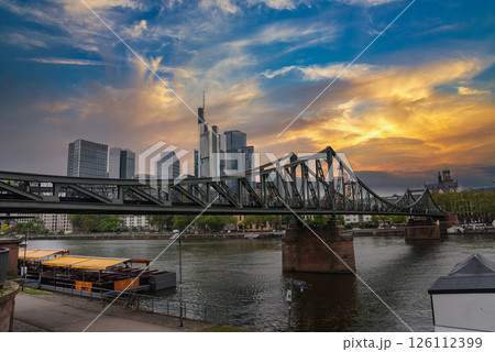 Eiserner Steg spans the Main River in Frankfurt, Germany, with a docked boat, historic cathedral spire, and modern skyscrapers under an overcast sky. Eiserner Steg spans the Main River in Frankfurt, Germany, with a docked boat, historic cathedral spire, and modern skyscrapers under an overcast sky. 126112399