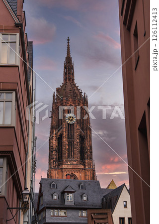 The Gothic style Frankfurt Cathedral stands tall with its intricate spire and clock, framed by modern and traditional buildings under a cloudy sky. 126112413