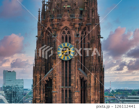 The Gothic style Frankfurt Cathedral tower stands out with intricate details and a gold accented clock, set against modern buildings and a pink blue sky. 126112441