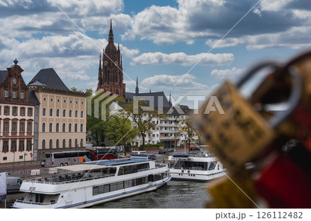Frankfurt Cathedral rises in the background, with boats along the river, historic European buildings, and love locks on a bridge in the foreground. 126112482