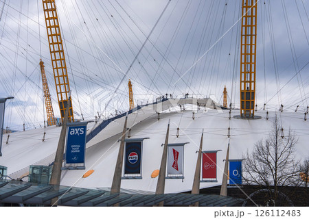 The O2 Arena in London features climbers on its white dome with yellow masts, brand banners in the foreground, and an overcast sky in the background. 126112483