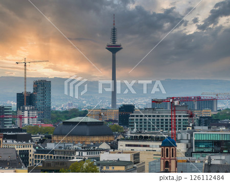 Aerial view of Frankfurt, Germany, featuring the Europaturm at sunset. Modern buildings, cranes, and rolling hills frame the urban skyline. 126112484