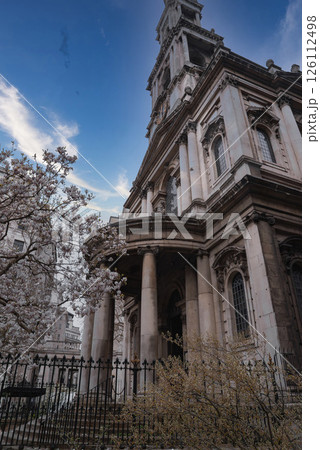 A historic London building featuring tall columns, arched windows, and stone detailing, surrounded by blooming trees under a partly cloudy sky. 126112498