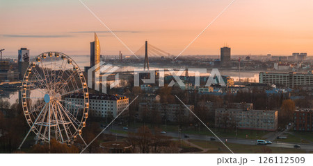Serene sunrise view of Riga, Latvia, featuring the Vansu Bridge, Z Towers, a large Ferris wheel, and the Daugava River reflecting warm light. Serene sunrise view of Riga, Latvia, featuring the Vansu Bridge, Z Towers, a large Ferris wheel, and the Daugava River reflecting warm light. 126112509