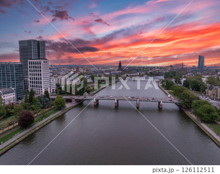 Aerial view of Frankfurt, Germany, at sunset. The Main River flows through the city, with a bridge, Eurotower, and Frankfurt Cathedral spire visible. 126112511