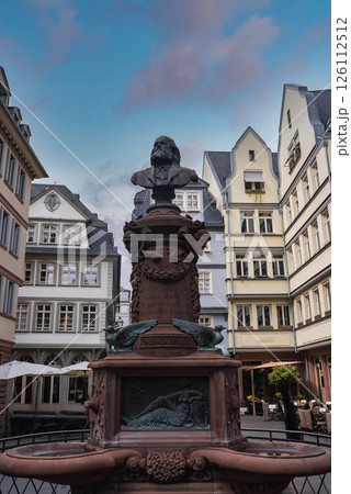 Statue of Friedrich Schiller atop an ornate fountain with intricate carvings, surrounded by European style buildings under a partly cloudy sky in Frankfurt. 126112512