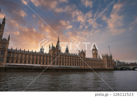 The Palace of Westminster and Elizabeth Tower with Big Ben at sunset, framed by a dramatic sky and the River Thames in the foreground. A crane is visible. 126112523