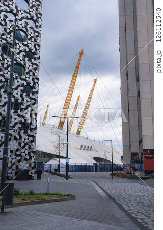 The O2 Arena's white dome and yellow masts stand out amid modern buildings, including one with a geometric facade, under a cloudy London sky. 126112540