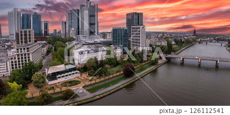 Aerial view of Frankfurt, Germany, at sunset featuring the Commerzbank Tower, Main River, a bridge, riverside greenery, and Frankfurt Cathedral spire. 126112541