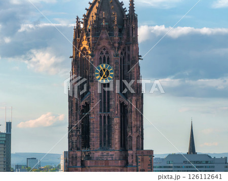 The Gothic style tower of Frankfurt Cathedral with intricate details, a gold accented clock, and modern buildings in the background under a cloudy sky. The Gothic style tower of Frankfurt Cathedral with intricate details, a gold accented clock, and modern buildings in the background under a cloudy sky. 126112641
