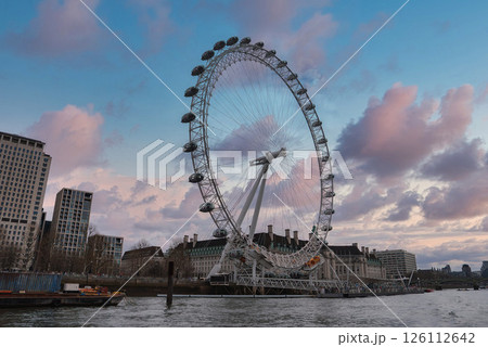 The London Eye stands along the River Thames with a vibrant pink and blue sky. Modern and historic buildings surround the landmark in this urban scene. The London Eye stands along the River Thames with a vibrant pink and blue sky. Modern and historic buildings surround the landmark in this urban scene. 126112642