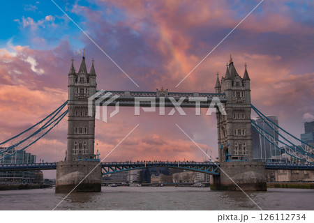 Tower Bridge spans the River Thames in London, featuring intricate towers, suspension cables, and a colorful sky. Modern buildings surround the historic landmark. 126112734