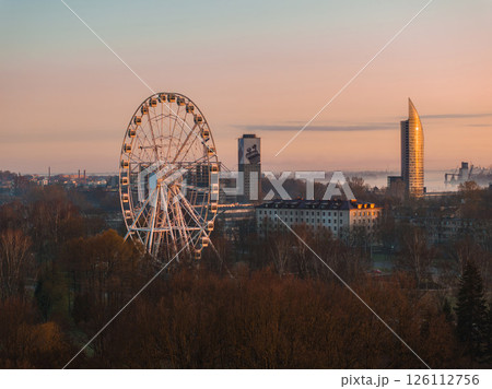 Serene morning scene in Riga, Latvia, featuring a Ferris wheel, Z Towers, bare trees, and mist over the Daugava River under soft golden light. Serene morning scene in Riga, Latvia, featuring a Ferris wheel, Z Towers, bare trees, and mist over the Daugava River under soft golden light. 126112756