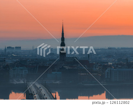 Serene sunrise over Riga, Latvia, featuring St. Peter's Church spire, a bridge over the Daugava River, and reflections in calm water with morning mist. Serene sunrise over Riga, Latvia, featuring St. Peter's Church spire, a bridge over the Daugava River, and reflections in calm water with morning mist. 126112802