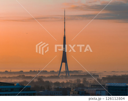 Serene morning scene in Riga, Latvia, featuring the iconic Riga TV Tower with its tripod design, light mist, and city rooftops under an orange sunrise. Serene morning scene in Riga, Latvia, featuring the iconic Riga TV Tower with its tripod design, light mist, and city rooftops under an orange sunrise. 126112804