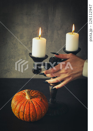 Female hand with sharp black nails touches vintage candlestick with burning candles, small pumpkin next to on the table, vertical image. 126112947