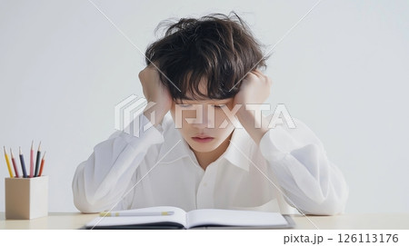 asian young boy sitting at a desk with his head in his hands. He is wearing a white collared shirt and appears to be stressed or frustrated	 126113176