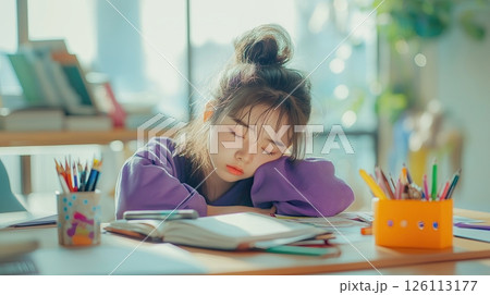 asian young girl sitting at a desk with her head resting on a book. She is wearing a purple shirt and has her hair tied up in a messy bun asian young girl sitting at a desk with her head resting on a book. She is wearing a purple shirt and has her hair tied up in a messy bun 126113177