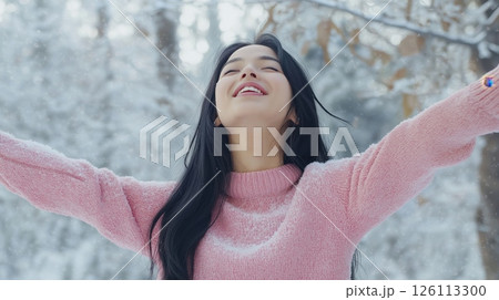 asian young woman standing in a snowy forest with her arms stretched out wide. She is wearing a pink sweater and has long black hair	 126113300
