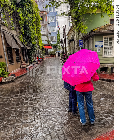 Couple holding pink umbrella strolls along a wet cobblestone street with greenery. High quality photo 126113554