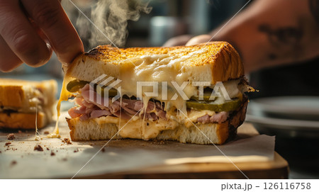 Hot Cuban sandwich being pulled apart with melted cheese strings and steam, showing layers of ham, pickles, and mustard in a warm bistro setting with golden light 126116758