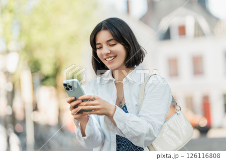 Mid-trip check-in with friends, multiracial Asian Latina Middle Eastern young woman with trendy short hair smiling and using phone in a European street 126116808