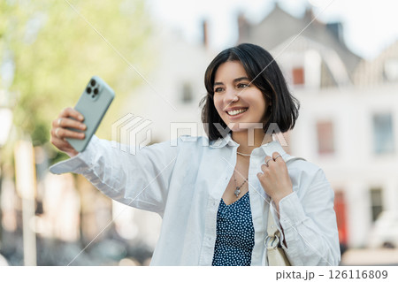 Replying to group chat updates, modern young woman with a fresh haircut and diverse background pauses mid-walk in a European city to take a selfie 126116809