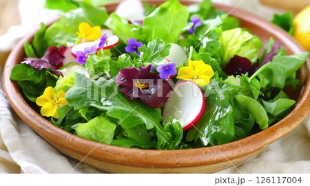 Fresh mixed greens with edible flowers and sliced radish on ceramic plate in soft daylight. Citrus dressing adds glistening highlights for an elegant editorial food shot. 126117004