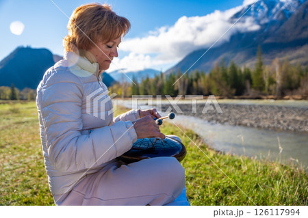 Woman playing a steel tongue drum in nature with a mountain river and Altai landscape in the background. Concept of harmony, mindfulness, and connection with nature 126117994