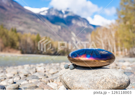 Steel tongue drum resting on a stone by the river in the Altai mountains. Concept of elemental harmony, raw sound energy, and nature as a sacred space for music and silence 126118003