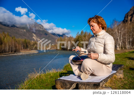 Woman playing a steel tongue drum in nature with a mountain river and Altai landscape in the background. Concept of harmony, mindfulness, and connection with nature 126118004