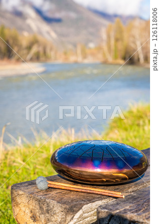 Steel tongue drum and mallets resting on a wooden bench with mountains, river, and forest in the background. Concept of sacred stillness, sound meditation, and harmony with nature 126118006