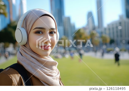 Young woman wearing headphones and a hijab smiles confidently in a vibrant urban park, with modern skyscrapers and greenery creating a lively atmosphere of city life 126118495