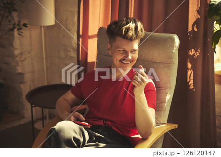 Man in red shirt sits comfortably in chair smiling and holding red smartphone. Rest at home in living room. Man in red shirt sits comfortably in chair smiling and holding red smartphone. Rest at home in living room. 126120137
