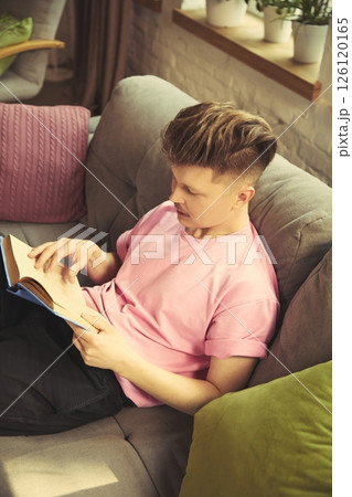 Man in pink shirt lying on sofa and reading book near large window in sunlight in cozy living room. Man in pink shirt lying on sofa and reading book near large window in sunlight in cozy living room. 126120165
