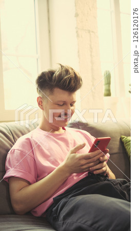 Happy man in pink shirt smiling, using smartphone, sitting on couch in cozy living room at home in daylight. 126120176