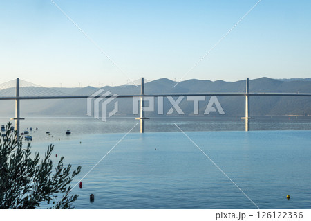 The Peljesac Bridge to Peljesac peninsula in Croatia, Europe. Suspension bridge spans tranquil water with mountains in the background, creating a serene coastal landscape with clear sky above. 126122336