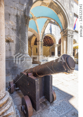 Korcula town in island of Korcula in Adriatic sea in Croatia, Europe. Historic cannon displayed in a courtyard with stone arches. Features a weathered barrel and wooden base, adding to the antiquity. 126122341